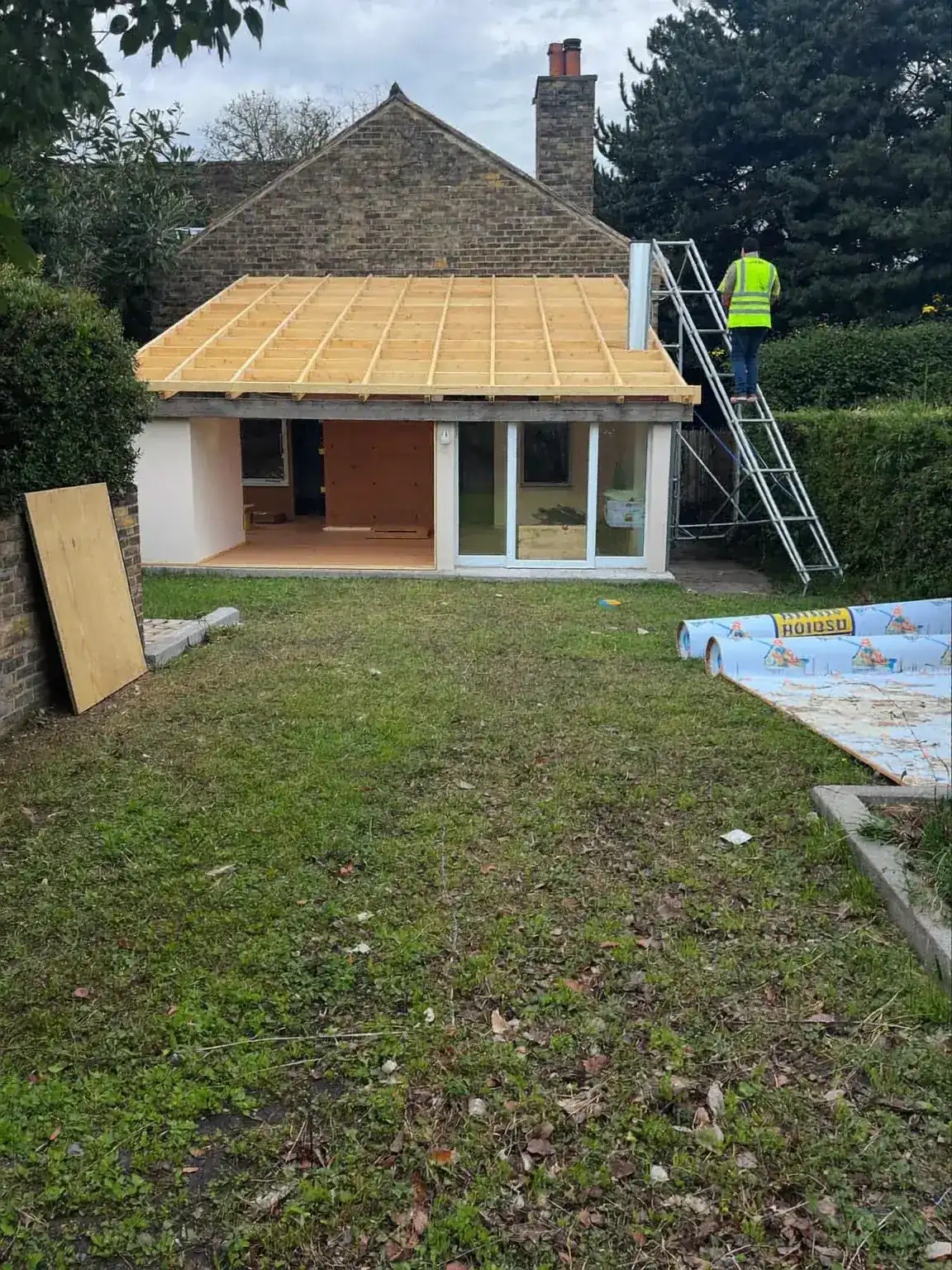 Timber roof joists fitted over a single-storey rear extension, with a ladder and plywood sheet in the garden