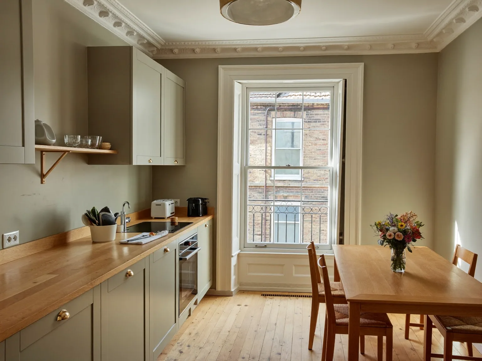 Sage-green shaker kitchen with oak worktops and a dining table beneath a tall sash window in a period room