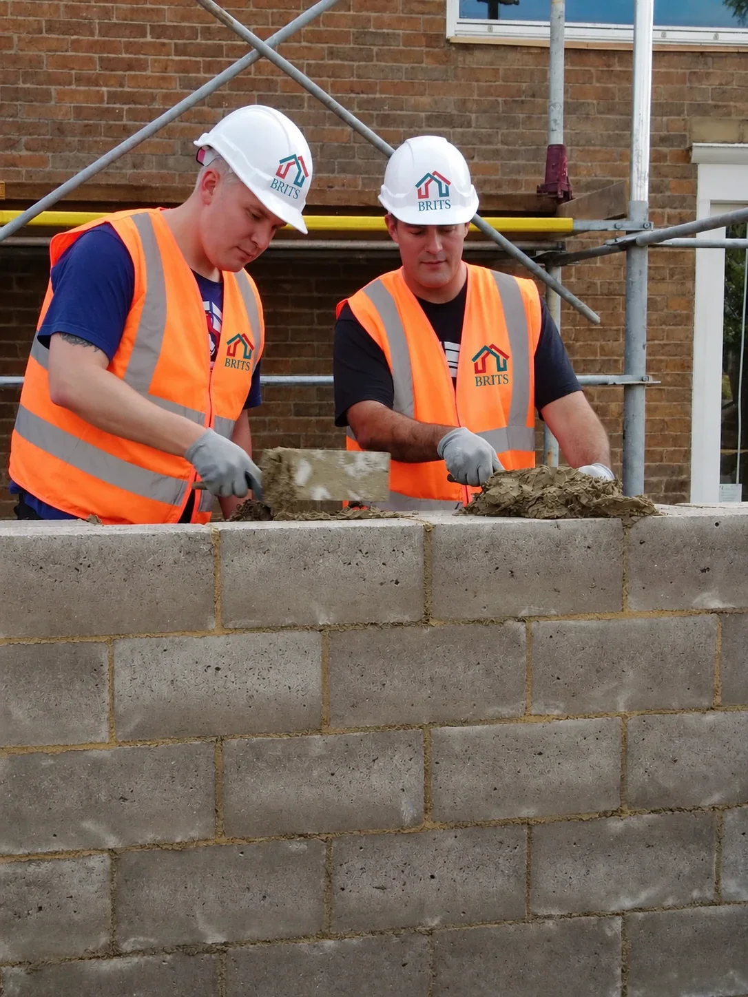 Two builders in Brits-branded hard hats and hi-vis laying a concrete-block wall