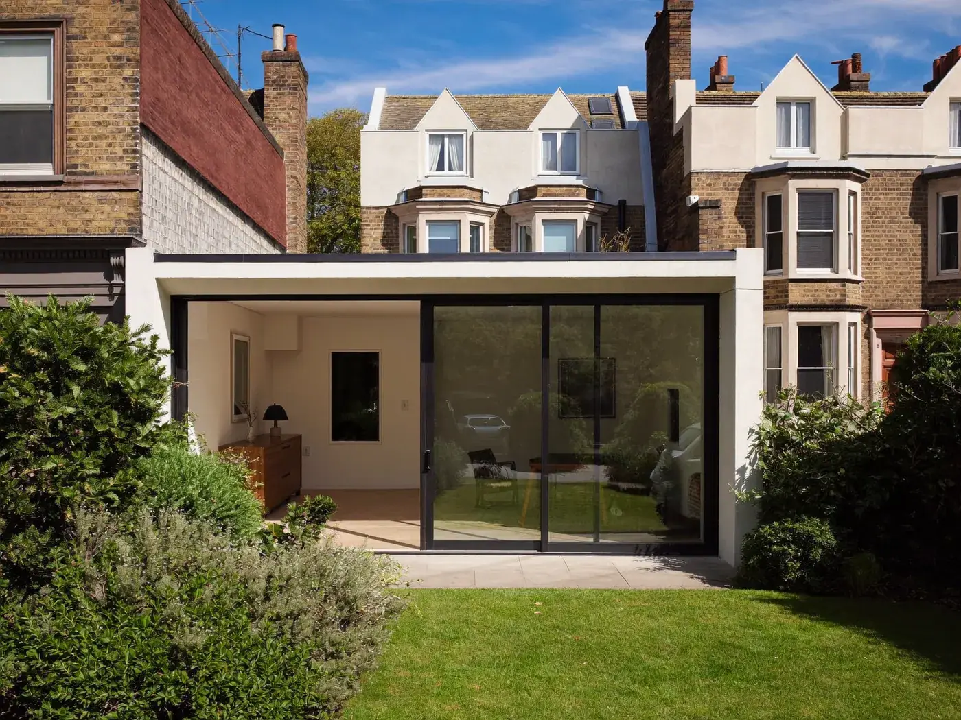 White-rendered rear extension with dark sliding glass doors below a tall brick terrace, with a lawn in front