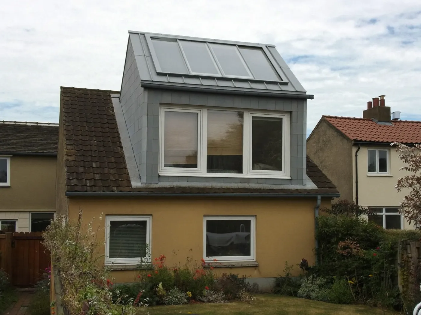 Grey-clad box dormer with roof windows on the rear roof of a yellow-rendered house, with white windows below
