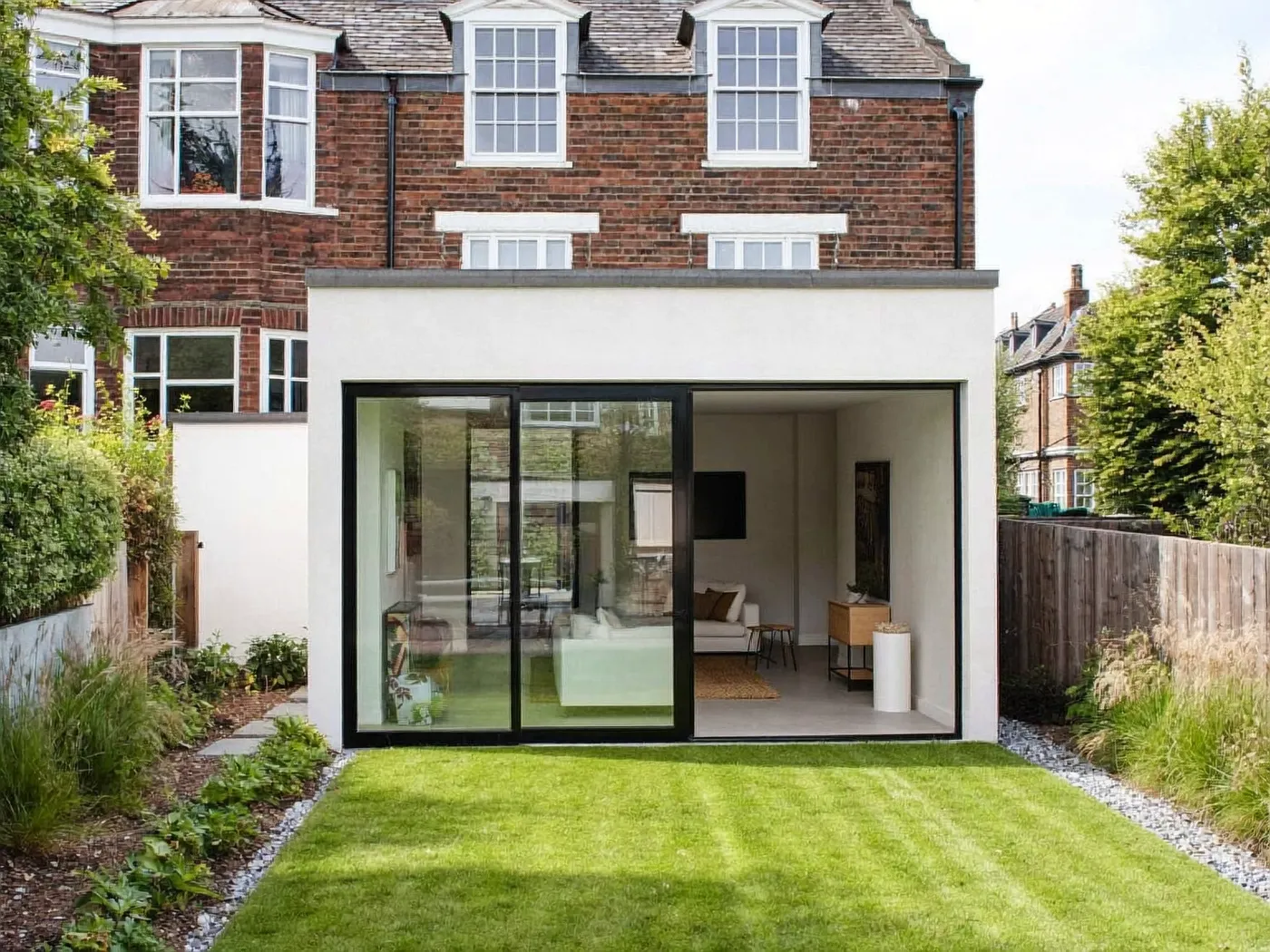 White-rendered rear extension with large dark-framed sliding glass doors below a red-brick terrace, with a lawn