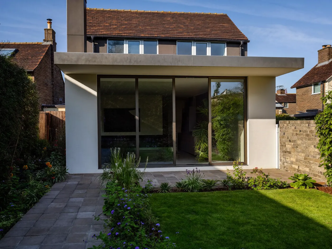 White-rendered rear extension with a flat roof overhang and sliding glass doors, with a paved path